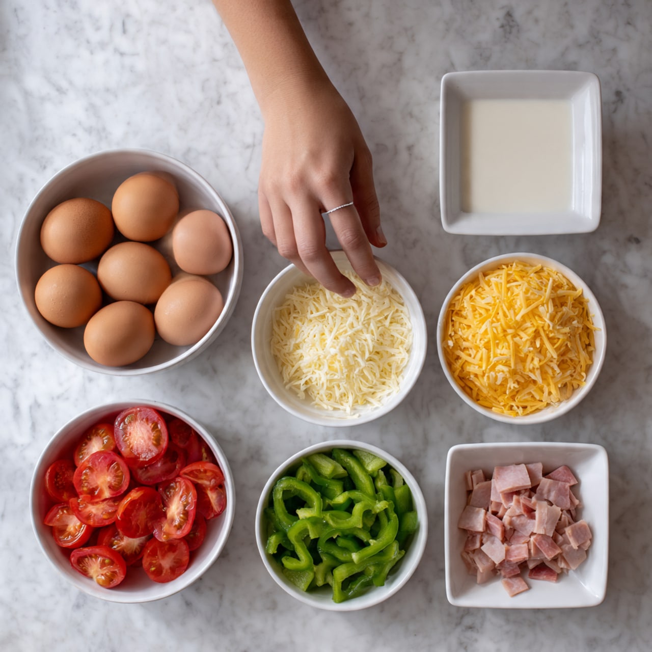 The image shows six white bowls arranged on a white marbled surface. From the top left, there is a round bowl filled with six brown eggs. To its right, a square bowl holds white milk, and next to it on the right, another square bowl contains grated yellow cheese. Below the eggs, a round bowl is filled with sliced red tomatoes. In the center bottom, a round bowl holds chopped green bell peppers. On the bottom right, a square bowl contains small pieces of sliced ham. A woman's hand enters from the left bottom corner, slightly touching the bowl of sliced tomatoes. photo taken with an iphone --ar 4:5 --v 7