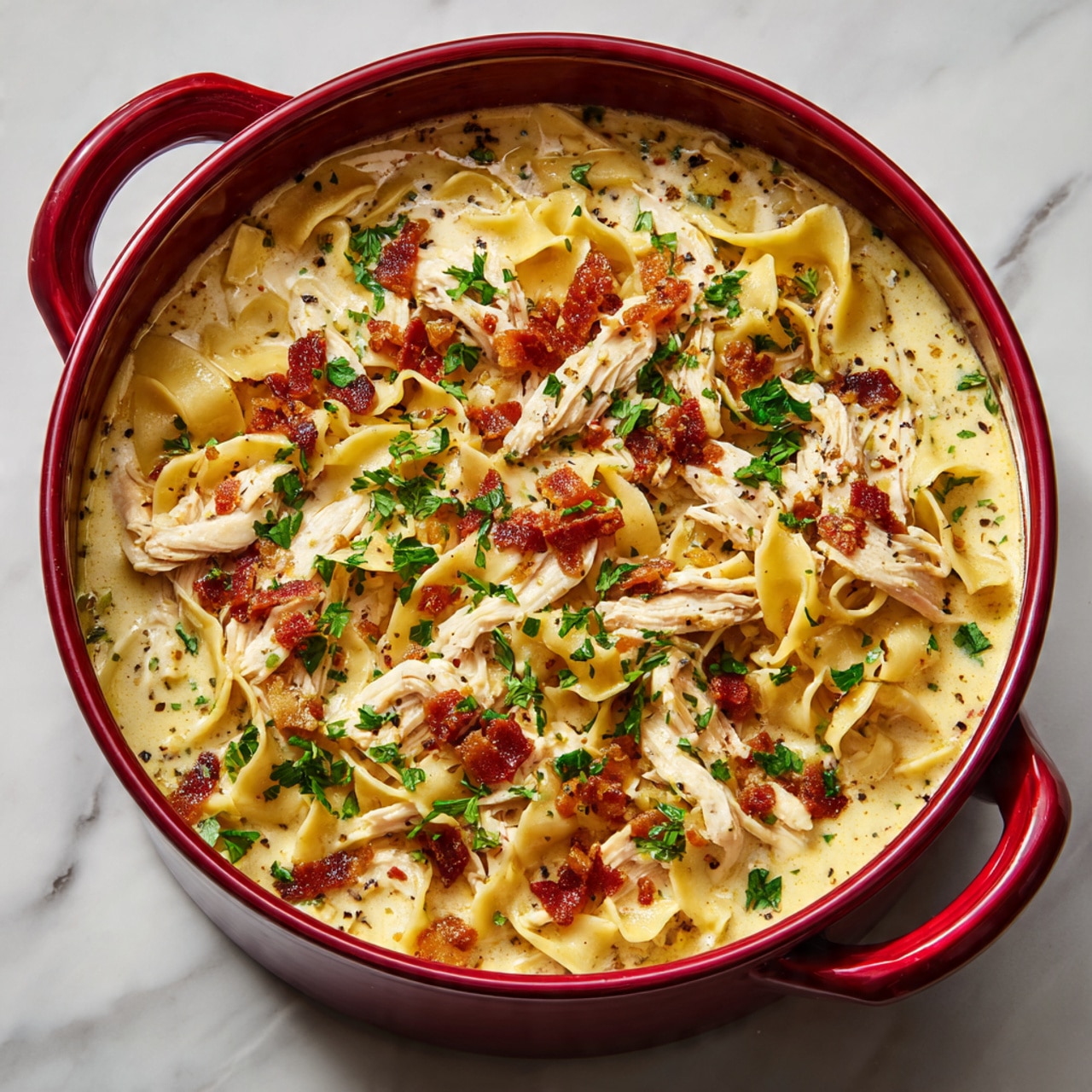 The image shows a deep red pot filled with creamy soup that has wide egg noodles as the bottom layer, covering the entire pot. On top of this, shredded white chicken pieces are scattered evenly. There is melted yellow cheese drizzled across the top, adding a rich texture. Crisp, small brown bacon bits are spread over the chicken and noodles, and fresh green parsley leaves are sprinkled over everything. The broth is thick and pale yellow with small specks of black pepper visible. The pot is placed on a white marbled surface. photo taken with an iphone --ar 4:5 --v 7