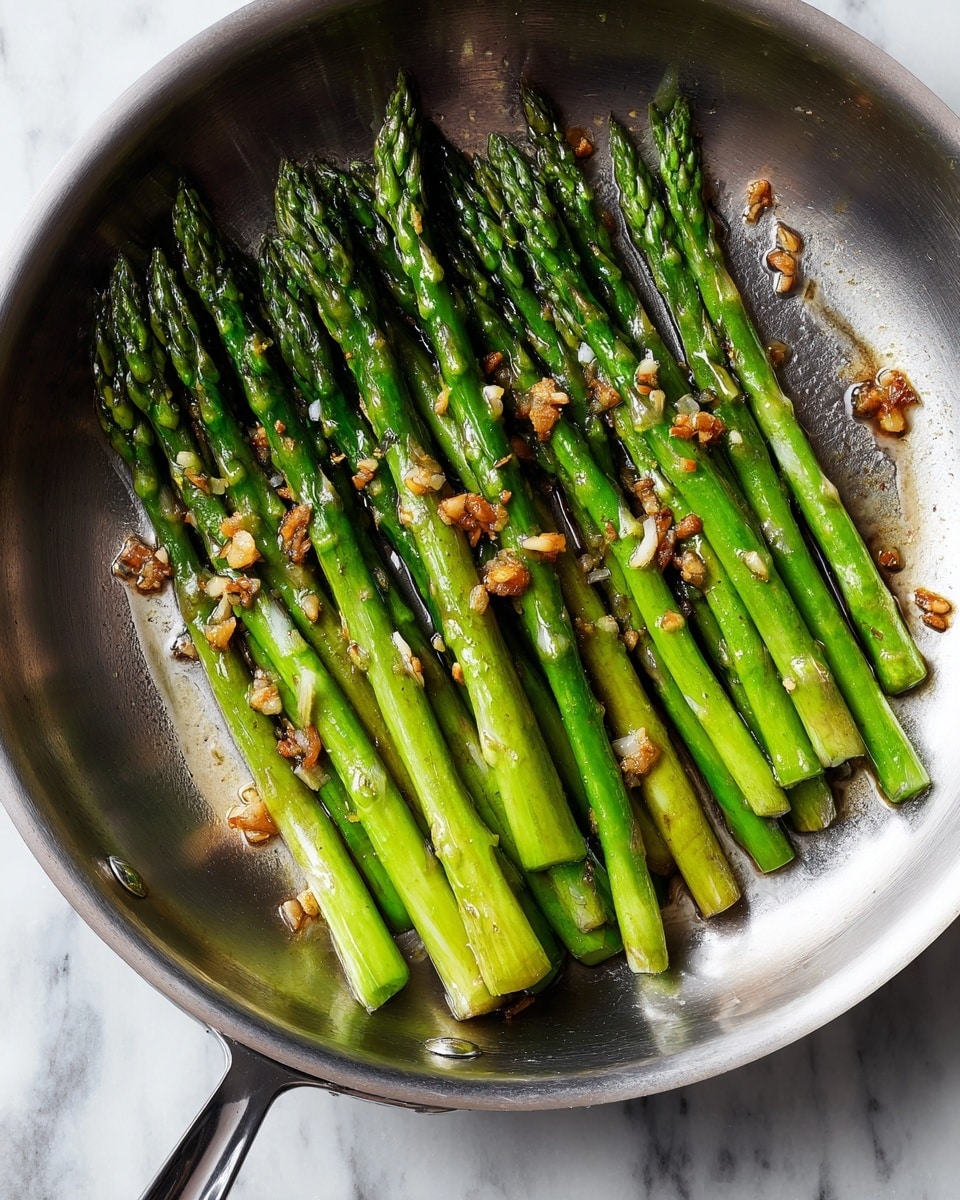 A stainless steel pan with many whole asparagus stalks aligned side by side, bright green in color with some toasted parts, topped with small pieces of light brown garlic bits scattered evenly across them, sitting on a white marbled surface. The asparagus has a glossy look from oil and the pan handle is visible, angled to the lower right. photo taken with an iphone --ar 4:5 --v 7