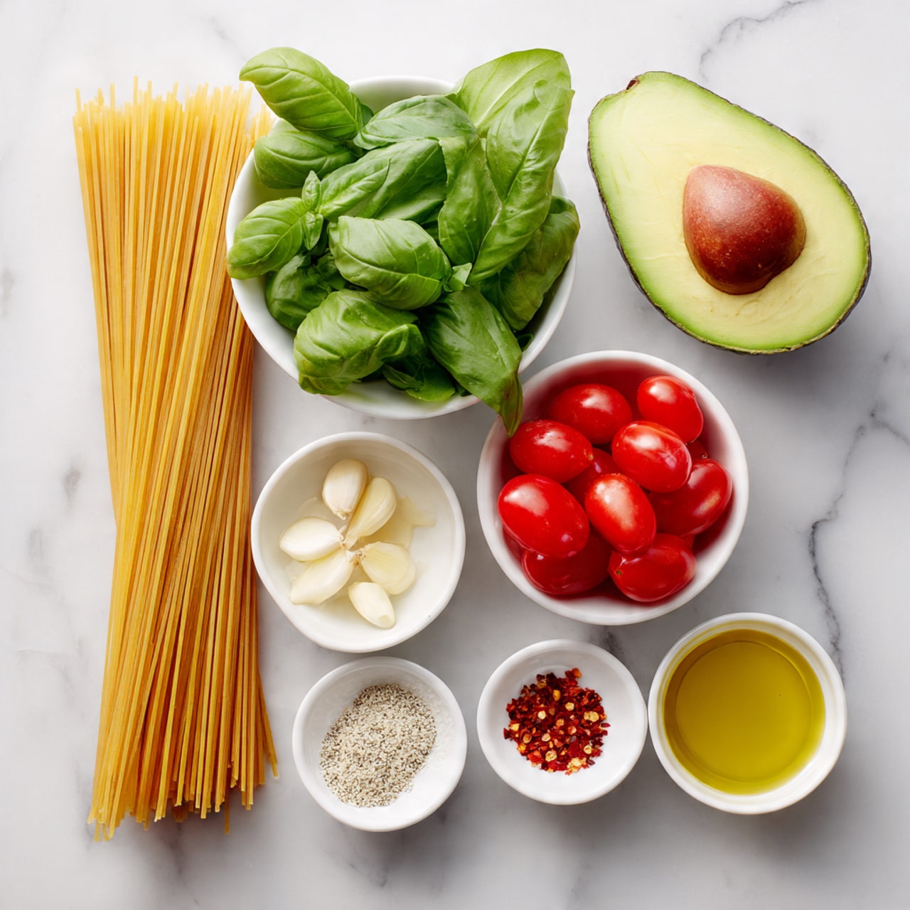 The image shows ingredients neatly placed on a white marbled surface. On the left is a bundle of uncooked spaghetti pasta, vertical with a smooth golden color. At the top right corner, there is a halved avocado with a bright green inside and a large brown seed in one half. Below the avocado is a white bowl filled with fresh green basil leaves. To the right of the basil, two white bowls contain halved red cherry tomatoes with juicy textures. Towards the bottom center, small white bowls hold olive oil with a shiny yellow-green surface, sliced garlic with white, thin pieces, coarse salt with a grainy white texture, and red chili flakes showing small dark red bits. The arrangement is clean and minimal. photo taken with an iphone --ar 4:5 --v 7