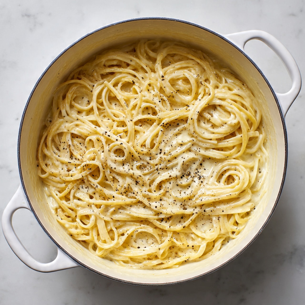 A white round pot filled with creamy pasta noodles covered in a thick light beige sauce with visible black pepper specks. The pasta strands are long, flat, and tangled, filling the pot evenly. The sauce has a smooth, glossy texture, evenly coating the noodles with some small bubbles and specks. The pot sits on a white marbled surface. photo taken with an iphone --ar 4:5 --v 7