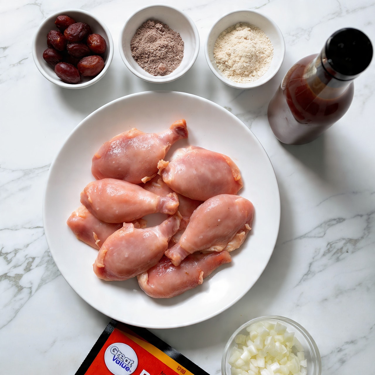 The image shows a white plate on a white marbled surface with several raw chicken pieces arranged neatly, showcasing skin and pink flesh textures. Above the plate, there are three small white bowls in a row: the left bowl has dark brown dates with a smooth, shiny texture; the middle bowl contains different light to dark brown powders and granules in three sections; the right bowl holds finely chopped white onions with a moist texture. To the right of the plate is a bottle of reddish barbecue sauce with a dark cap, standing upright. Below the plate, there is a black and yellow packet labeled