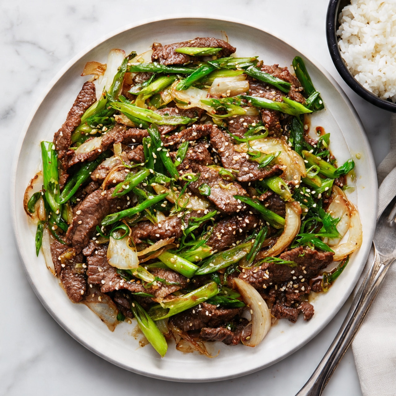 A large white plate holds cooked thin slices of brown beef mixed with sautéed light brown onion strips and sprinkled with small white sesame seeds. Bright green sliced scallions are scattered on top, adding color contrast. The plate is set on a white marbled surface, with a black bowl containing white rice visible on the right side. A silver fork lies at the bottom edge of the plate. photo taken with an iphone --ar 4:5 --v 7