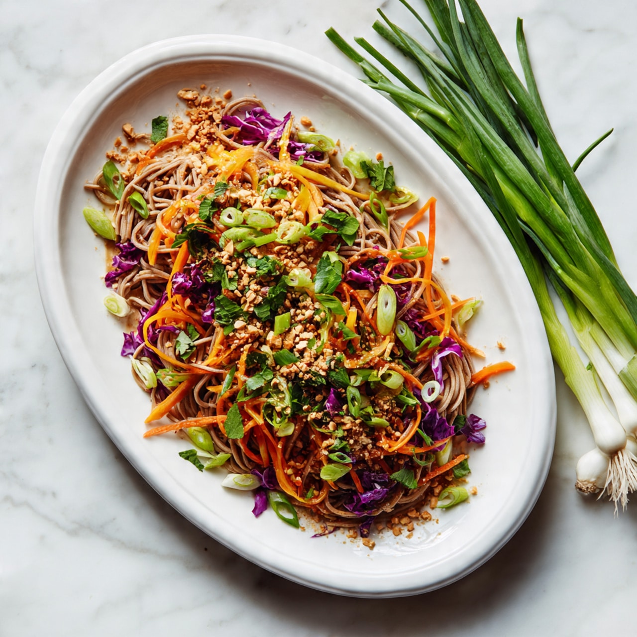 On a white long oval plate, there are several layers of light brown soba noodles forming the base layer with a slightly rough texture. On top, thin carrot strips in bright orange, fine purple cabbage shreds, and chopped green herbs are scattered evenly, adding vibrant colors. A sprinkling of crushed nuts or seeds is spread gently over the vegetables, and thinly sliced green onions are placed around the edges, giving a fresh look. The plate rests on a white marbled surface with some green onions nearby. Photo taken with an iphone --ar 4:5 --v 7