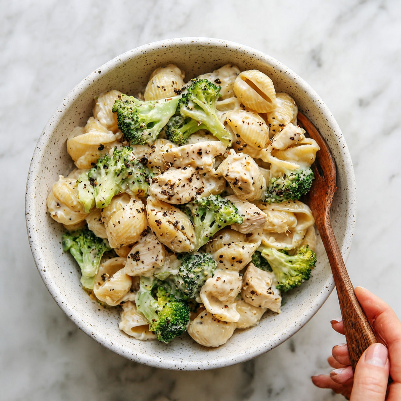 A white speckled bowl filled with creamy pasta, featuring light green broccoli florets and small pieces of light brown chicken mixed evenly throughout. The pasta is coated in a smooth, off-white sauce with visible black pepper specks sprinkled on top. A woman's hand holding a wooden fork is reaching into the bowl from the top right. The bowl is placed on a white marbled surface. photo taken with an iphone --ar 4:5 --v 7
