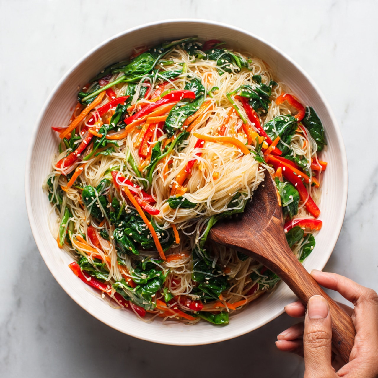 A round white bowl filled with a colorful noodle salad that has about four layers: the bottom layer is thin translucent noodles with a shiny texture, the next layer shows thin slices of red bell peppers and shredded carrots mixed evenly, followed by bright green spinach leaves spread throughout, and on top there are small sesame seeds sprinkled. A wooden spoon with a woman's hand holding it is lifting some noodles from the bowl. The surface under the bowl is white marble. Photo taken with an iphone --ar 4:5 --v 7