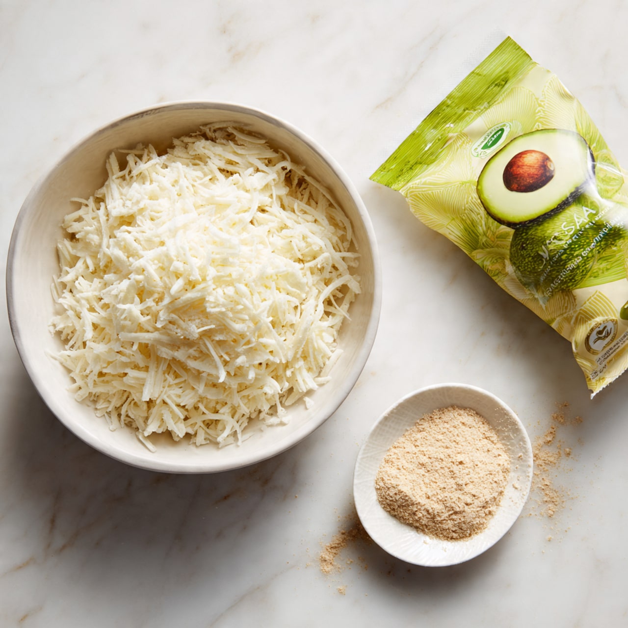 The image shows a white bowl filled with shredded white cheese placed on a white marbled surface. Next to it, there is a small white plate holding a light brown powder. Above the small plate, a partially opened green and yellow snack or food package with an avocado image on it lies on the white marbled surface. The scene is bright and clean. photo taken with an iphone --ar 4:5 --v 7