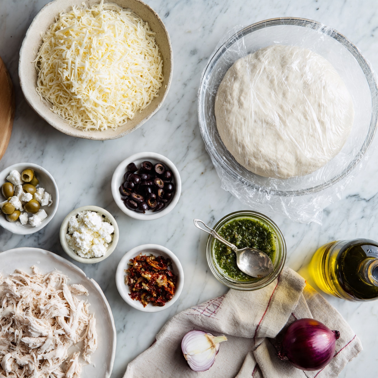 A top-down view of various pizza ingredients arranged on a white marbled surface, featuring a large clear glass bowl with white dough covered partially with plastic wrap on the right side, a white plate with shredded chicken in the bottom left, and a white bowl filled with shredded cheese in the upper left; nearby are three small white ramekins containing black olives, white crumbled cheese, and chopped sun-dried tomatoes, while a small clear container with green pesto sauce and a silver spoon sits in the middle, a halved red onion rests on a folded beige and white cloth near a dark green bottle of extra virgin olive oil on the right; everything is clean and neatly placed, well-lit with natural light, photo taken with an iphone --ar 4:5 --v 7