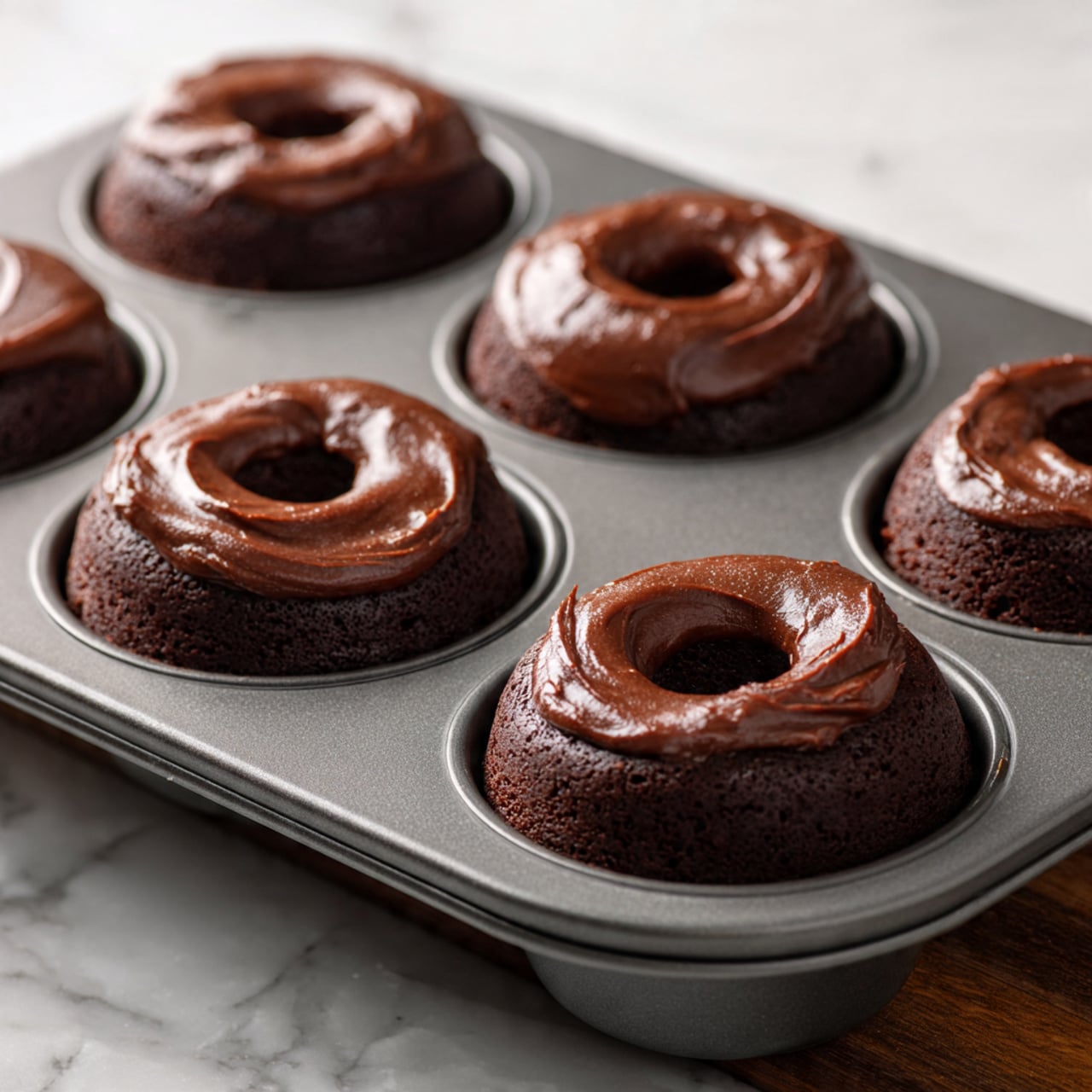 The image shows a dark gray metal donut baking tray filled with six portions of thick chocolate batter, each sitting in a round hollow with a raised center post. The chocolate batter is rich and smooth with a slightly glossy surface, dark brown in color, and fills each donut mold almost to the top. The table beneath the tray has a wooden look but should be imagined as a white marbled texture. The tray is slightly angled, giving a close-up view of the front three donut batter portions with clear detail on the texture of the batter and mold shapes. photo taken with an iphone --ar 4:5 --v 7