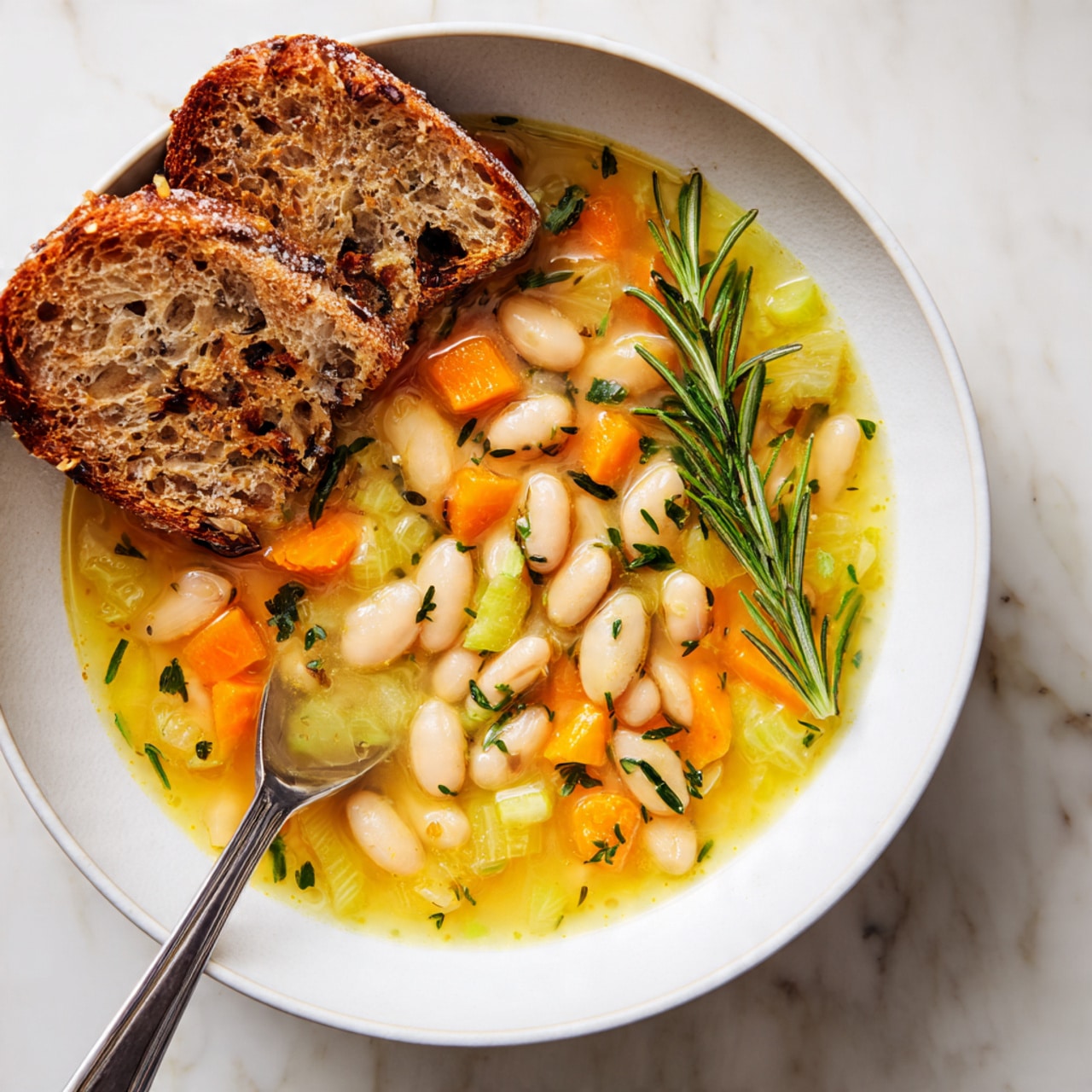 A white bowl filled with light yellow broth soup containing large white beans, small orange carrot cubes, and pale green celery pieces, all mixed with finely chopped herbs floating on surface. On the right side near the edge, a sprig of fresh rosemary rests in the soup. Three pieces of toasted multigrain bread stand vertically on the left side slightly inside the bowl. A metal spoon rests partially submerged in the soup on the bottom left. The bowl sits on a white marbled texture surface. Photo taken with an iphone --ar 4:5 --v 7