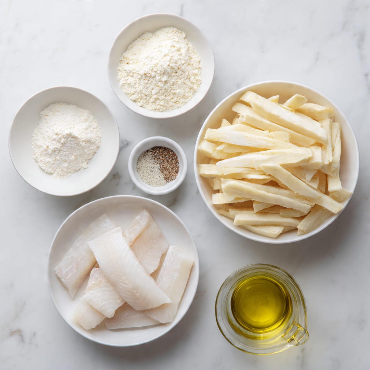 The image shows six white bowls and a transparent jug all arranged on a white marble surface. The largest bowl on the bottom left holds three pieces of light, raw white fish with smooth texture. To the right of it, another large bowl contains many pale yellow potato sticks that look firm and freshly cut. Above these, there are four smaller bowls: one with white flour, another with a white powder (possibly cornstarch), one with salt and pepper mixed, and the last with a white fine powder (likely baking powder). On the bottom right, a transparent jug contains a golden-yellow liquid that looks like oil. The setup is clean and well organized, photographed with soft, natural light. photo taken with an iphone --ar 4:5 --v 7