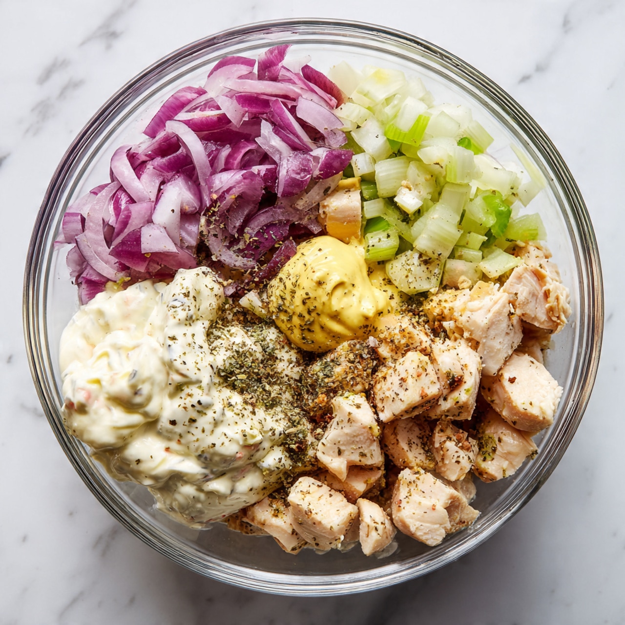 A clear glass bowl sits on a white marbled surface filled with layers of ingredients for a salad. At the bottom right, there are chunks of pale beige cooked chicken. To the left of the chicken, a thick dollop of creamy white mayonnaise rests beside a small scoop of yellow mustard. At the top, thin slices of purple-red onion are arranged next to diced green celery pieces. A sprinkle of black ground pepper is visible over the chicken and onions. The ingredients are all unmixed, showing distinct layers and colors. Photo taken with an iphone --ar 4:5 --v 7
