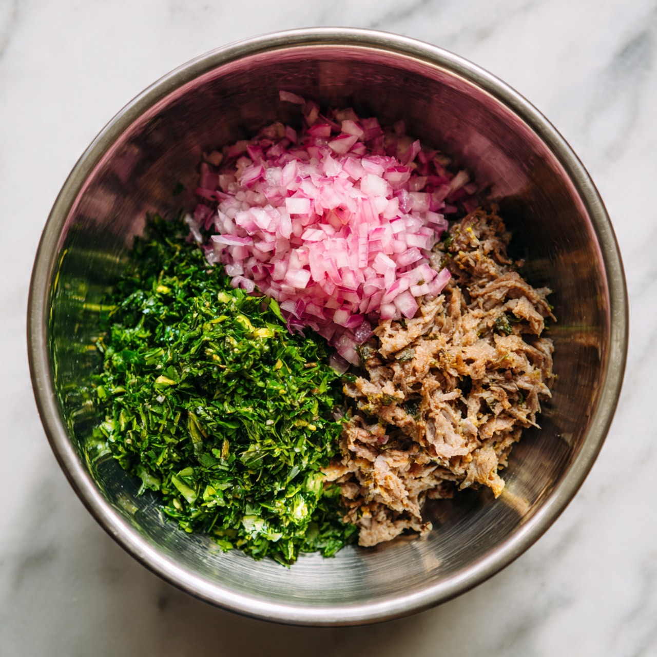 Inside a shiny metal bowl, three main layers sit next to each other. On the left is a pile of bright green chopped herbs with a fresh texture. To the right of the herbs is a mound of finely chopped pink onions, showing small bits and a soft texture. At the bottom right sits a portion of cooked, crumbly brown meat had small crisp bits. The three layers contrast well inside the bowl, which reflects light softly. The image is set against a white marbled texture. photo taken with an iphone --ar 4:5 --v 7