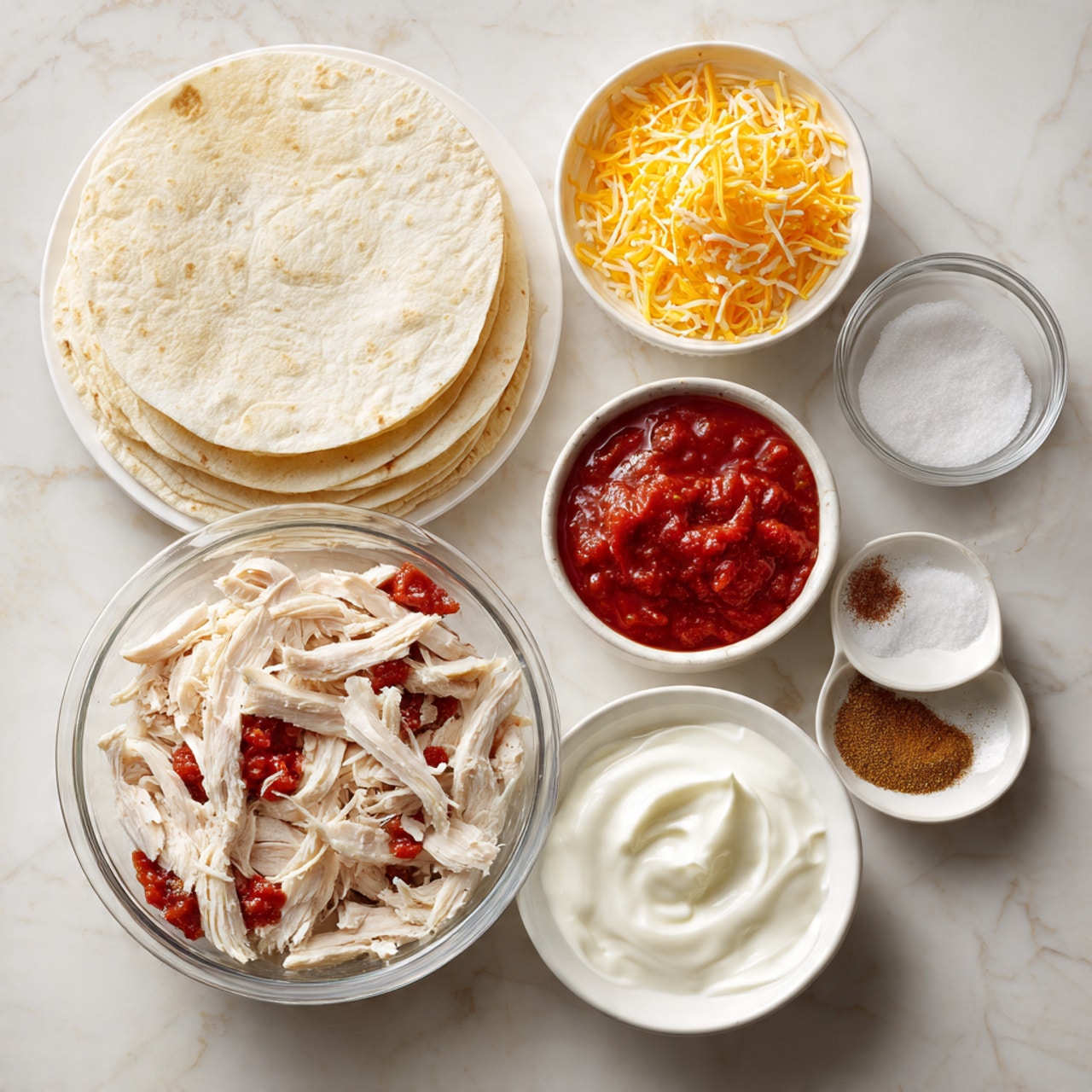 A top view of cooking ingredients arranged on a white marbled surface: a clear glass bowl in the center contains layers of finely chopped red tomatoes with juice, white sour cream, and several spices including a light brown powder and a darker brown powder; above it, a white bowl filled with finely shredded orange cheese; to the right, a small white bowl with a small amount of white salt; below the glass bowl, a white bowl holds shredded light brown cooked chicken; and to the bottom left, a white plate is stacked with several round, light beige tortillas. photo taken with an iphone --ar 4:5 --v 7