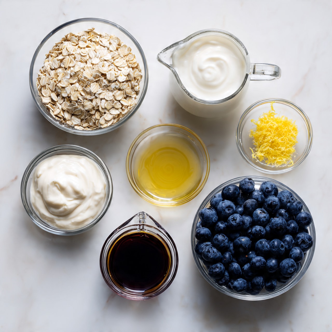 The image shows seven small clear glass bowls and a clear glass measuring jug arranged on a white marbled surface. Starting from the top left is a bowl filled with light beige rolled oats, next to it is the measuring jug filled with white milk. To the right is a bowl of thick white yogurt. Below the oats is a small bowl with white salt, beside it is a small bowl with bright yellow lemon zest, and next to that is a small bowl with pale yellow lemon juice. In the middle bottom is a small glass measuring jug filled with dark amber vanilla extract. To the bottom right is a bowl full of fresh, plump blueberries in dark blue and purple shades. The overall look is clean and simple, with the clear bowls showing the different textures and colors clearly. Photo taken with an iphone --ar 4:5 --v 7
