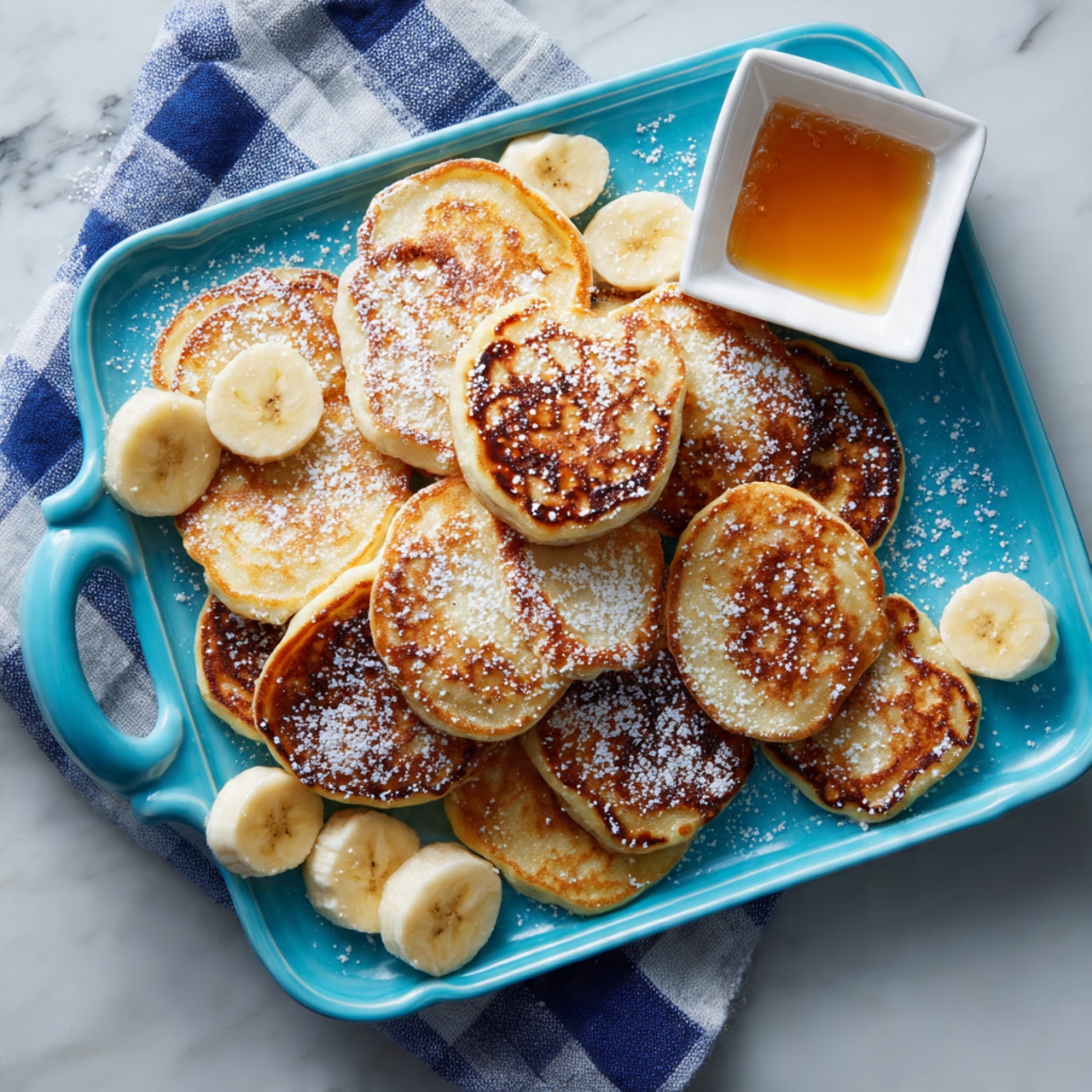 A light blue rectangular plate with a handle holds about twenty small oval pancakes stacked loosely in the center. The pancakes are golden brown with some darker spots and have a soft, fluffy texture. There is a dusting of white powdered sugar spread unevenly over the pancakes. Around the edges of the plate, there are three large banana halves, peeled and placed lengthwise with a drizzle of honey on top. A small white square dish with golden syrup sits near the top right corner of the plate. The plate is set on a white marbled surface with a blue and white checkered cloth partially under it. photo taken with an iphone --ar 4:5 --v 7
