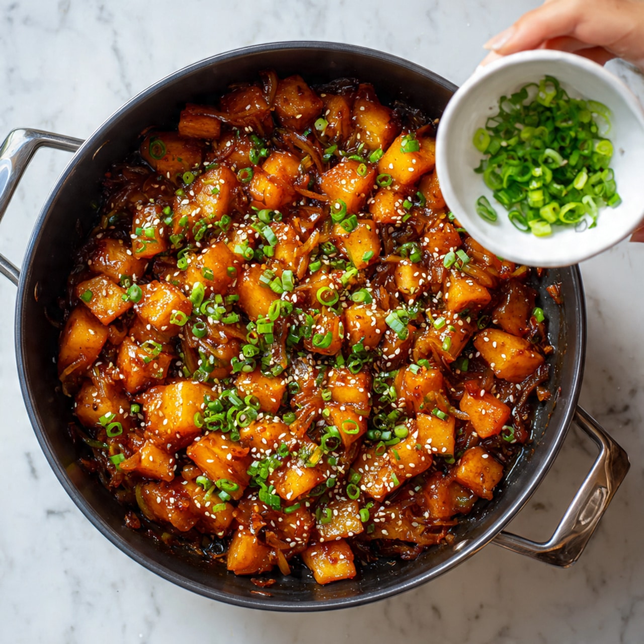 A close-up of a black pot filled with orange-brown cubed pieces of cooked potatoes and onions in a shiny dark sauce, topped with white sesame seeds and small green chopped scallions. The pot is on a white marbled surface. On the right, a woman's hand holds a small white bowl pouring extra chopped scallions into the pot. The colors are warm with shiny sauce reflections and small bits of green and white scattered evenly on top photo taken with an iphone --ar 4:5 --v 7