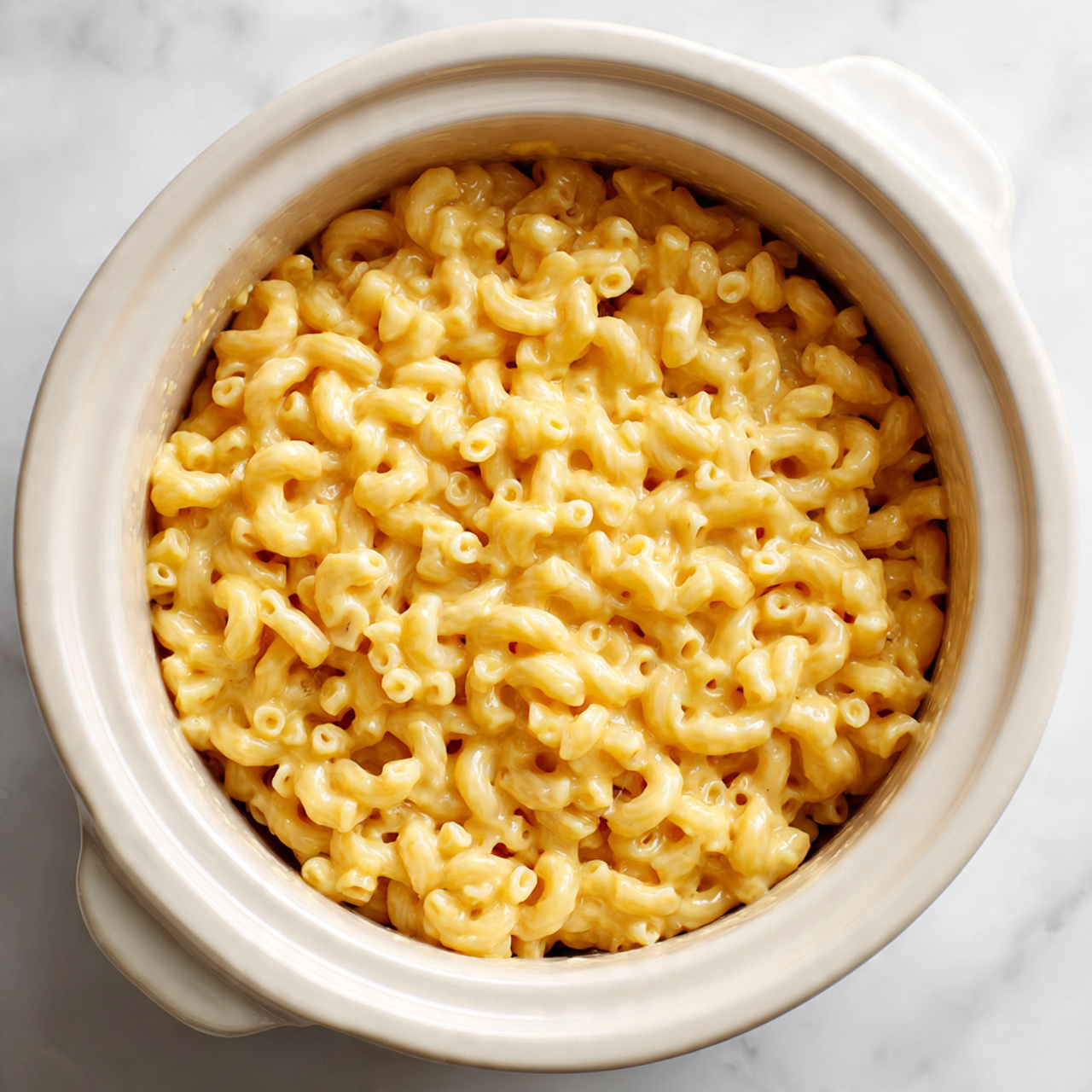 A close-up view of a white slow cooker filled with creamy macaroni and cheese. The macaroni noodles are curved and are coated in a smooth yellow cheese sauce, filling the pot evenly without any toppings or garnishes. The slow cooker rests on a white marbled surface. The lighting highlights the glossy texture of the cheese sauce on the noodles. Photo taken with an iphone --ar 4:5 --v 7
