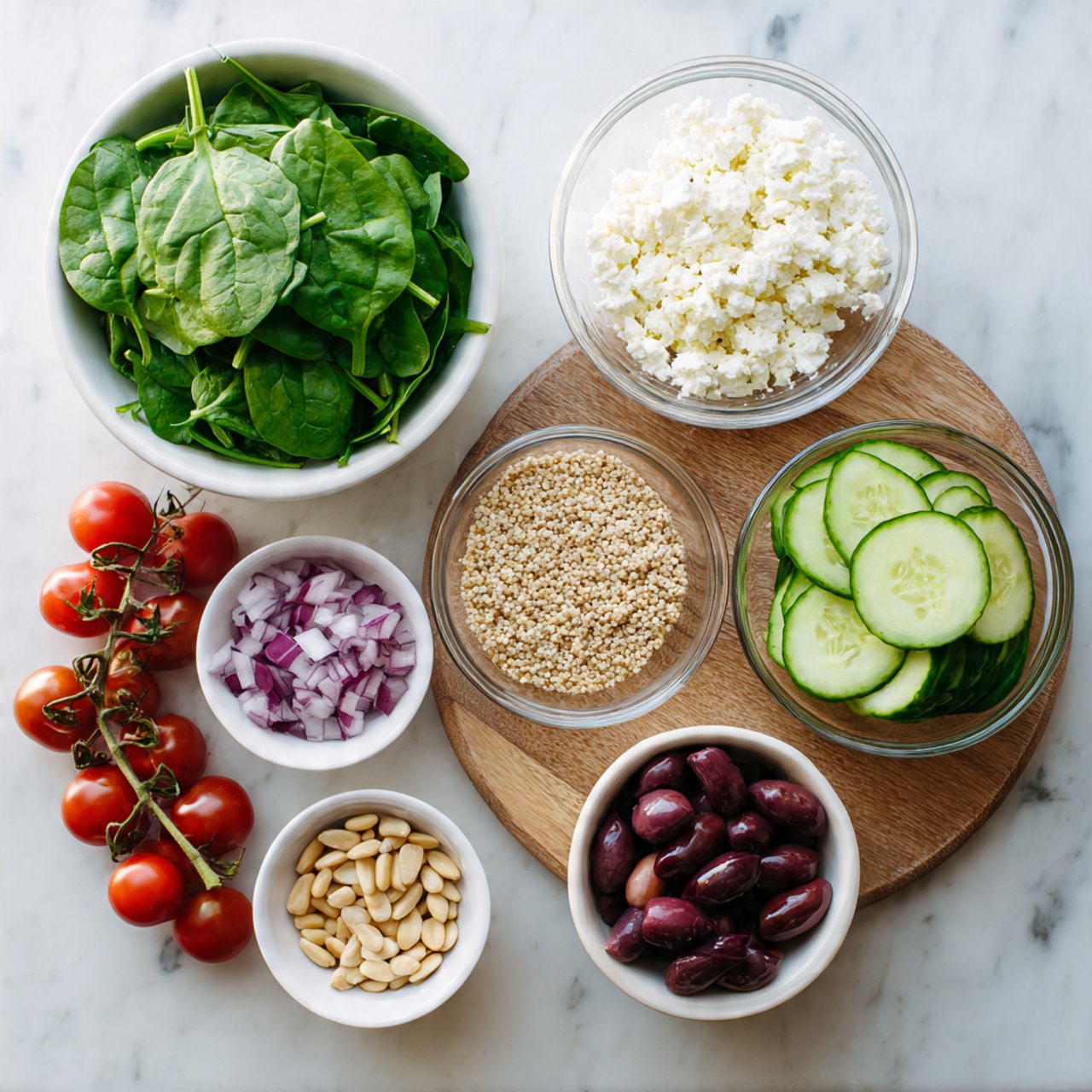 A white bowl filled with fresh green spinach leaves sits at the top left. Next to it on the right is a small clear glass bowl full of crumbled white feta cheese. Below the cheese is a clear bowl with uncooked light beige quinoa grains. Below the quinoa is another small clear bowl holding chopped red onions. To the left of the onions, near the bottom of the image, is a small white bowl filled with pale beige pine nuts. At the bottom left, a bunch of red cherry tomatoes on the vine lies across the surface. Above the tomatoes is a small pile of dark purple olives. In the center of the image is a round wooden board with sliced light green cucumbers stacked on top. The entire setup is on a white marbled surface. Photo taken with an iphone --ar 4:5 --v 7