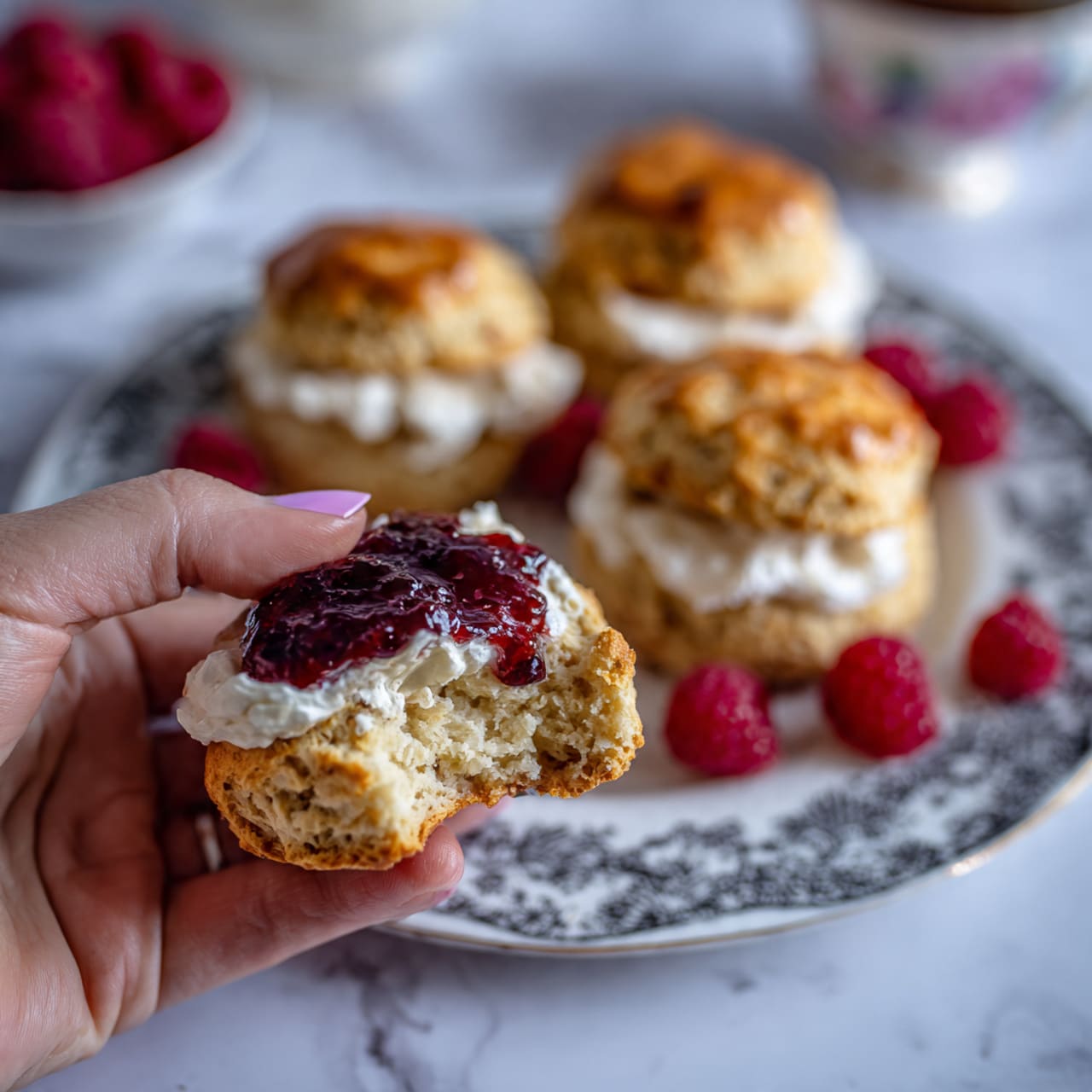 The image shows a woman's hand holding a bitten golden biscuit with a soft, slightly crumbly inside, topped with white cream and dark red jam. In the background, three whole biscuits with shiny golden tops are placed on a white plate featuring a detailed black pattern. Fresh red raspberries are scattered on the plate around the biscuits. The setting is on a white marbled surface. photo taken with an iphone --ar 4:5 --v 7
