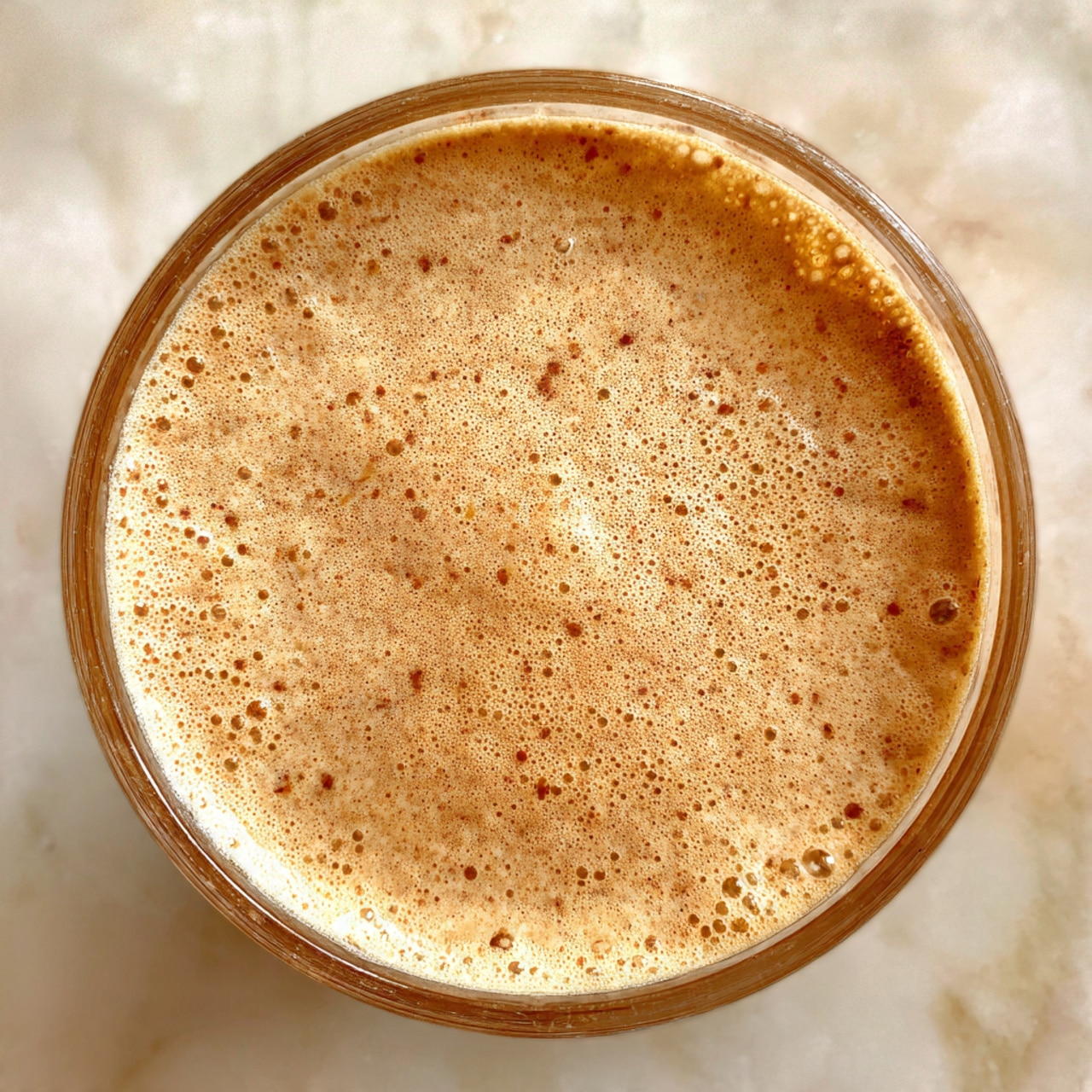 A clear blender container filled with a light brown creamy mixture that has tiny specks of darker brown throughout. The mixture has a smooth, slightly foamy texture with small bubbles on the surface, seen from above against a white marbled background. photo taken with an iphone --ar 4:5 --v 7