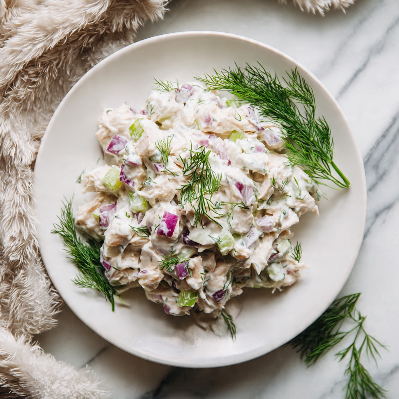 The image shows a white plate filled with creamy chicken salad. The salad has small pieces of chicken mixed with light cream-colored dressing, dotted with small bits of purple onion and green celery. Fresh dill sprigs are placed on top and around the salad, adding a touch of green. The plate sits on a white marbled surface with a fluffy, light fabric partially visible in the background. Photo taken with an iphone --ar 4:5 --v 7