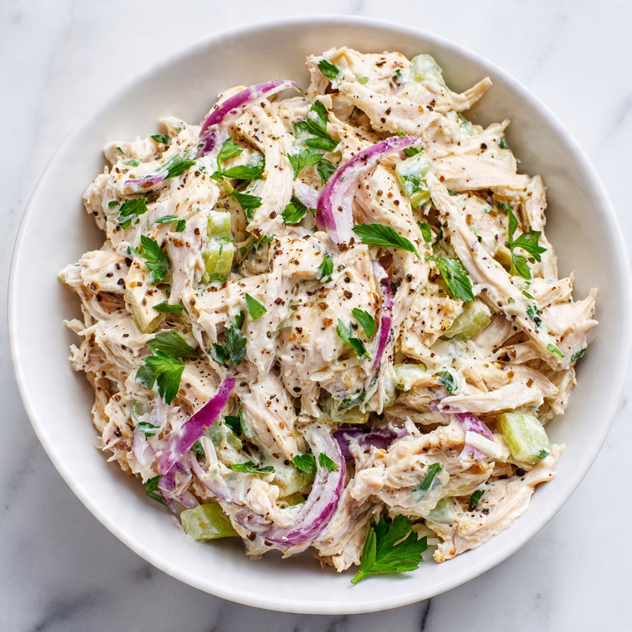 A white bowl filled with creamy chicken salad sits on a white marbled surface. The salad has three main layers: shredded light beige chicken mixed with a light creamy dressing, chunky pale green celery pieces, and thin slices of purple-red onion. Small green parsley leaves are scattered throughout and on top as garnish. The salad has visible black pepper sprinkled on it, adding a touch of dark contrast. In the background, out-of-focus green parsley adds more color to the scene. Photo taken with an iphone --ar 4:5 --v 7