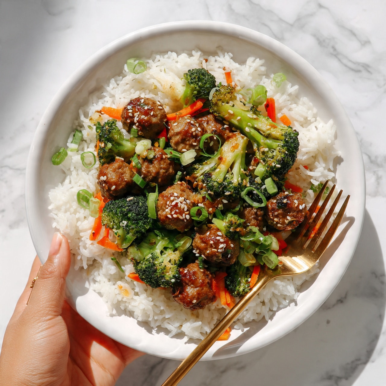 The image shows a plate of cooked white rice topped with a mix of small meatballs, broccoli florets, thin carrot strips, and chopped green onions, all coated in a light sauce with visible sesame seeds. The dish is served on a white plate placed on a white marbled surface. A golden fork is positioned at the bottom right part of the plate with a woman's hand holding the fork. The colors are bright, with the green vegetables, orange carrots, and brown meatballs contrasting nicely against the white rice and plate. The scene looks fresh and inviting, with natural lighting. photo taken with an iphone --ar 4:5 --v 7