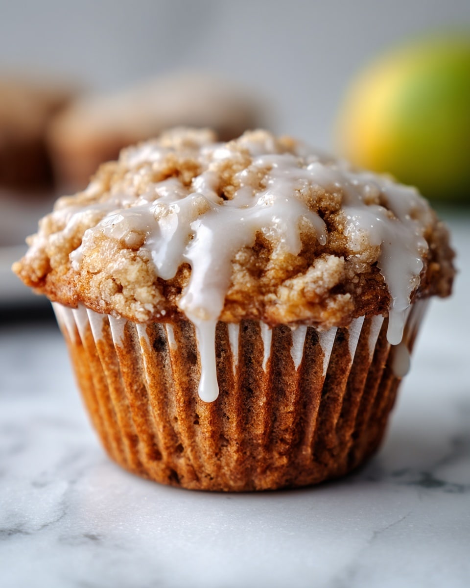 A close-up of a single muffin topped with a light drizzle of white icing that softly flows down its crumbly, golden-brown surface. The muffin has a rough and textured top with visible chunks inside the soft, warm brown base. It sits on a white paper liner with subtle creases, placed on a white marbled surface. In the blurred background, there is a hint of a large green shape, possibly fruit. The photo is bright with natural light, highlighting the muffin’s crackly texture and the smooth icing on top. photo taken with an iphone --ar 4:5 --v 7