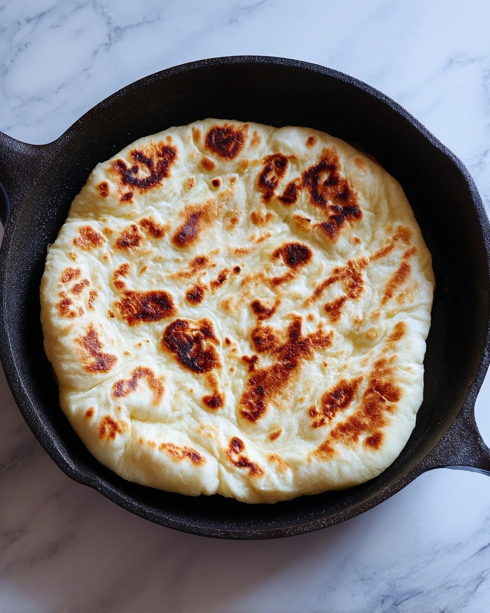 The image shows a single piece of puffed flatbread cooking in a black pan. The flatbread has a light beige color with small dark brown spots scattered on its surface, indicating areas where the bread has browned. Its texture looks soft and slightly fluffy, with a raised middle section giving it a swollen, pocket-like shape. The pan's dark surface contrasts with the pale bread, and the lighting highlights the bread's gentle curves and uneven puffiness. The scene is set against a white marbled texture. photo taken with an iphone --ar 4:5 --v 7