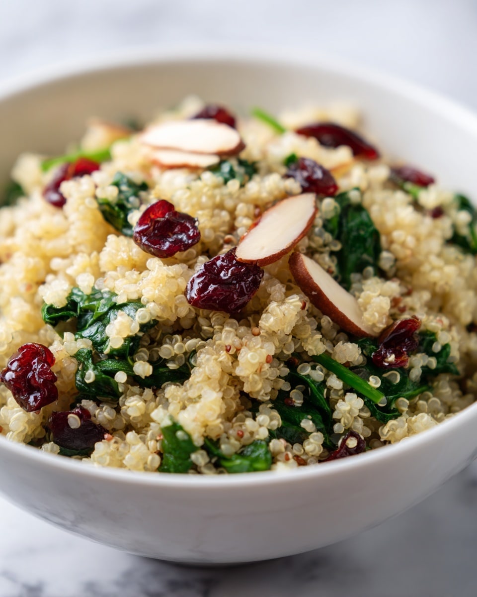 A close-up view of a white bowl filled with a mixed quinoa salad, showing three main layers: light fluffy quinoa grains with a pale yellow color form the base, topped with dark green leafy spinach pieces evenly spread throughout, and scattered on top are small bright red dried cranberries and pale beige almond slices adding contrast and texture. The salad's mix of colors and soft textures stands out against a white marbled surface. photo taken with an iphone --ar 4:5 --v 7