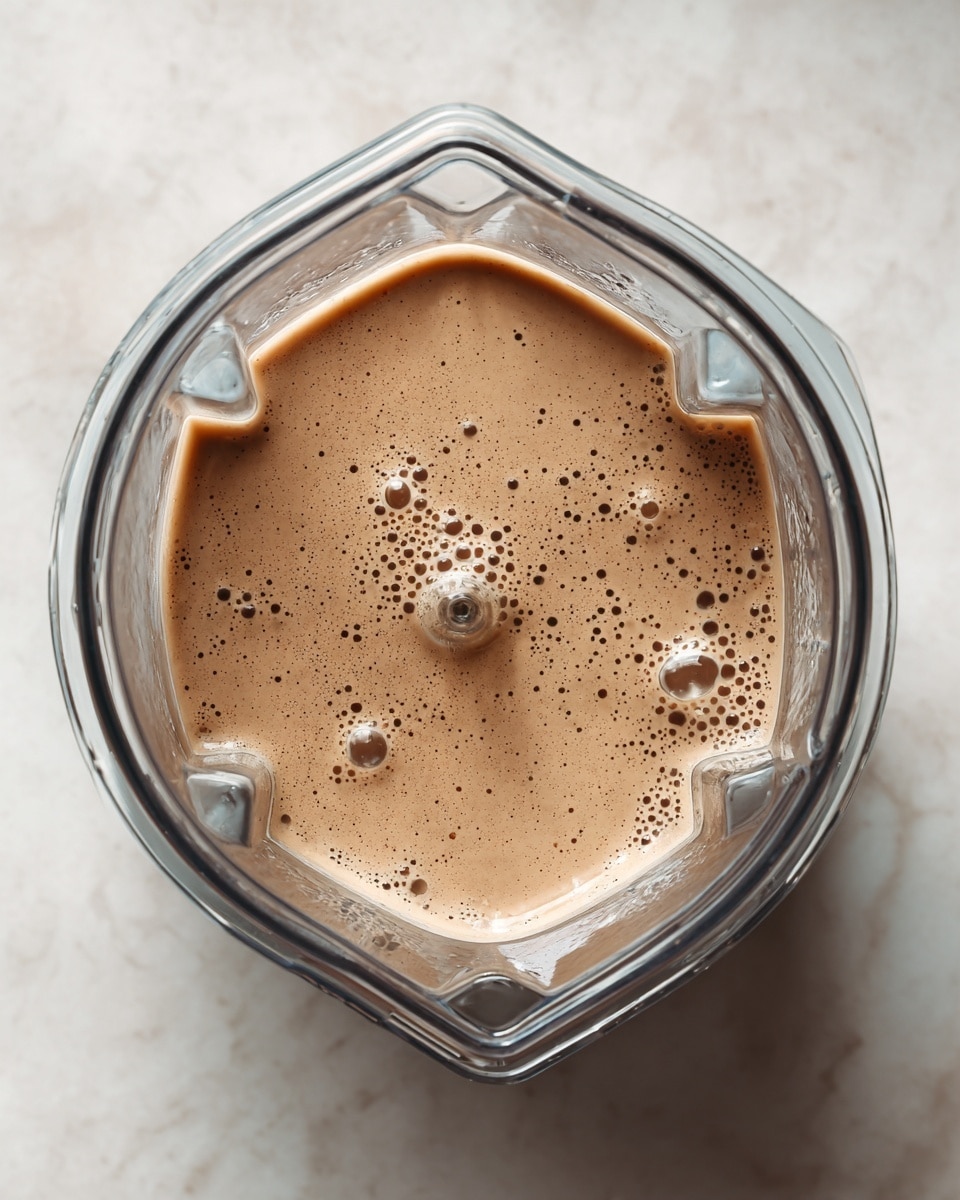 The image shows a clear blender jar viewed from above, filled with a smooth, light brown creamy liquid with tiny darker specks evenly spread inside. The liquid surface has several small bubbles, giving it a soft, frothy look. The blender jar is set on a white marbled surface and has a slightly glossy texture reflecting light, with defined edges and geometric shapes on the inside walls of the jar. Photo taken with an iphone --ar 4:5 --v 7