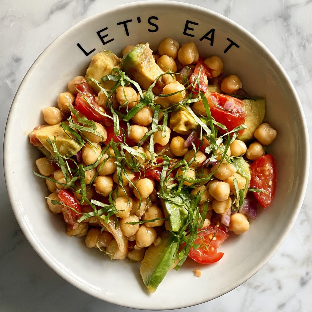 square image The image shows a white bowl filled with a chickpea salad. The base layer is made of light beige chickpeas that are round and plump. Scattered throughout are medium-sized pieces of green avocado and bright red tomato chunks, alongside small bits of purple onion for color contrast. Thin strips of fresh green basil leaves are spread evenly on top, giving the salad a fresh and vibrant look. The bowl is placed on a white marbled surface, with the words
