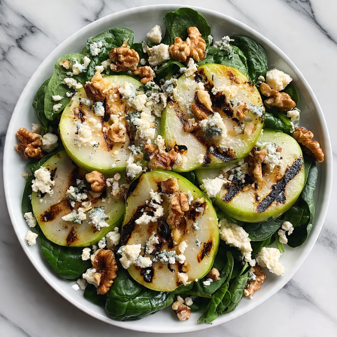 square image A white plate filled with a fresh salad showing about three layers: the bottom layer is dark green spinach leaves with a smooth texture, the middle layer has light green grilled pear slices with dark grill marks, and the top layer is scattered walnuts and crumbled cheese pieces with a creamy texture. The plate is placed on a white marbled surface, and the colors of the salad include deep green, light green, brown, and white. photo taken with an iphone --ar 4:5 --v 7square image