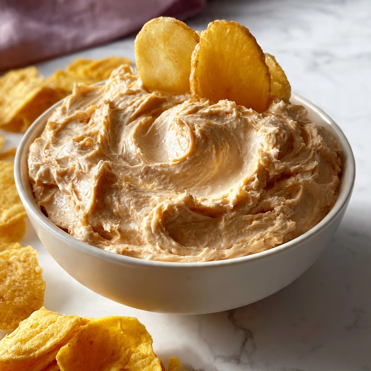 square image A white bowl filled with a creamy, swirled light brown dip, topped with a few potato chips standing upright in the dip. Around the bowl, there are scattered yellowish, thick tortilla chips. The bowl sits on a white marbled surface with a soft purple cloth slightly seen in the background. The texture of the dip is smooth but has visible swirls and a slight thick consistency. Photo taken with an iphone --ar 4:5 --v 7square imagesquare image