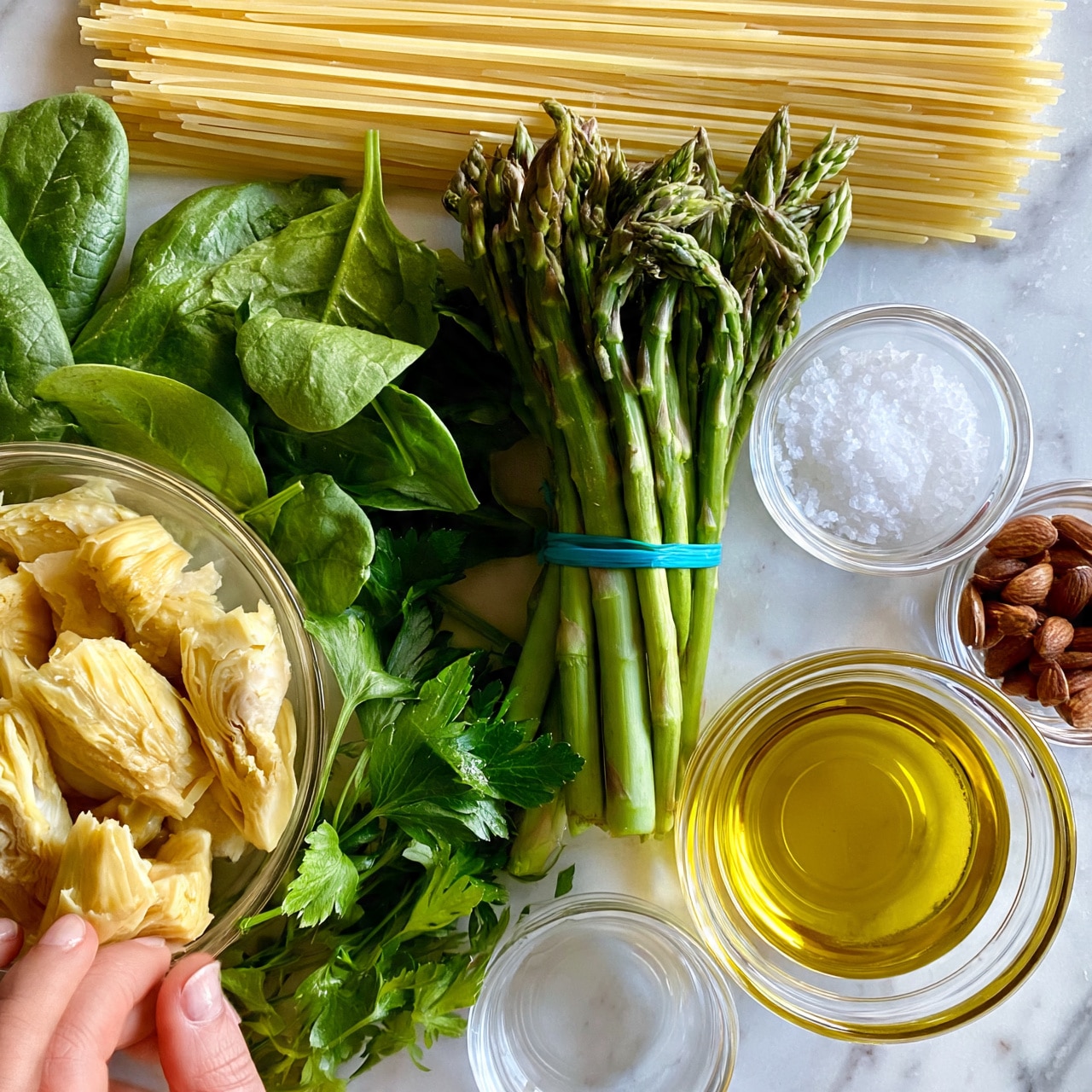 Asparagus Pesto Pasta Recipe 4 square image This image shows ingredients for cooking, placed on a white marbled surface. At the top is a neat bundle of raw spaghetti, pale yellow and straight. Below it, a bunch of green asparagus stands upright, tied with a blue band. To the left in a clear bowl, there are light yellow artichoke pieces with a slightly rough texture. Below and around the bowls are fresh green leaves of spinach and parsley, their textures leafy and vibrant. To the right, small clear glass bowls hold coarse white salt and a handful of brown almonds. Near the bottom center, a clear glass bowl contains golden-yellow olive oil that looks smooth and shiny. A woman's hand, partly shown, touches the artichoke bowl. photo taken with an iphone --ar 4:5 --v 7