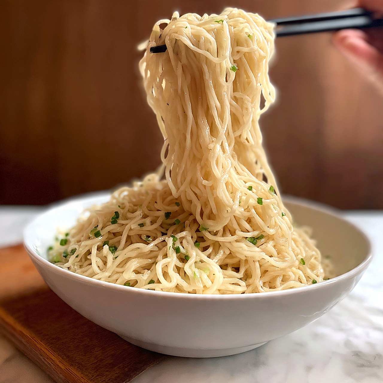square image A bowl of noodles in a white bowl is set on a white marbled surface with a wooden board nearby. The noodles are light beige with a slightly shiny, soft texture, piled high in the bowl. Green tiny chopped herbs or scallions are sprinkled on top, adding small green dots of color. A pair of black chopsticks held by a woman's hand lifts a large amount of noodles above the bowl, showing the noodles' long, thin strands twisting and hanging down smoothly. The background has a warm, blurred brown wooden wall, adding softness to the scene. photo taken with an iphone --ar 4:5 --v 7