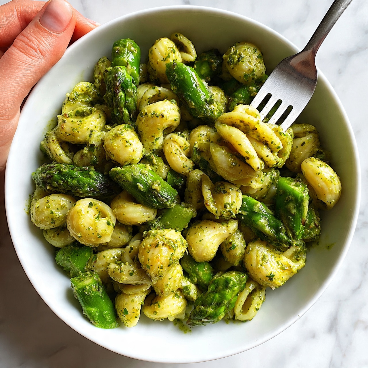 Asparagus Pesto Pasta Recipe 5 square image The image shows a white bowl filled with twisted pasta coated in green pesto sauce. The pasta is mixed with bright green asparagus pieces, cut into short lengths and scattered evenly through the dish. The green pesto clings to the pasta and vegetables, giving a rich, slightly chunky texture with visible bits of herbs. A silver fork held by a woman's hand is twirling some pasta inside the bowl. The bowl sits on a white marbled surface. photo taken with an iphone --ar 4:5 --v 7