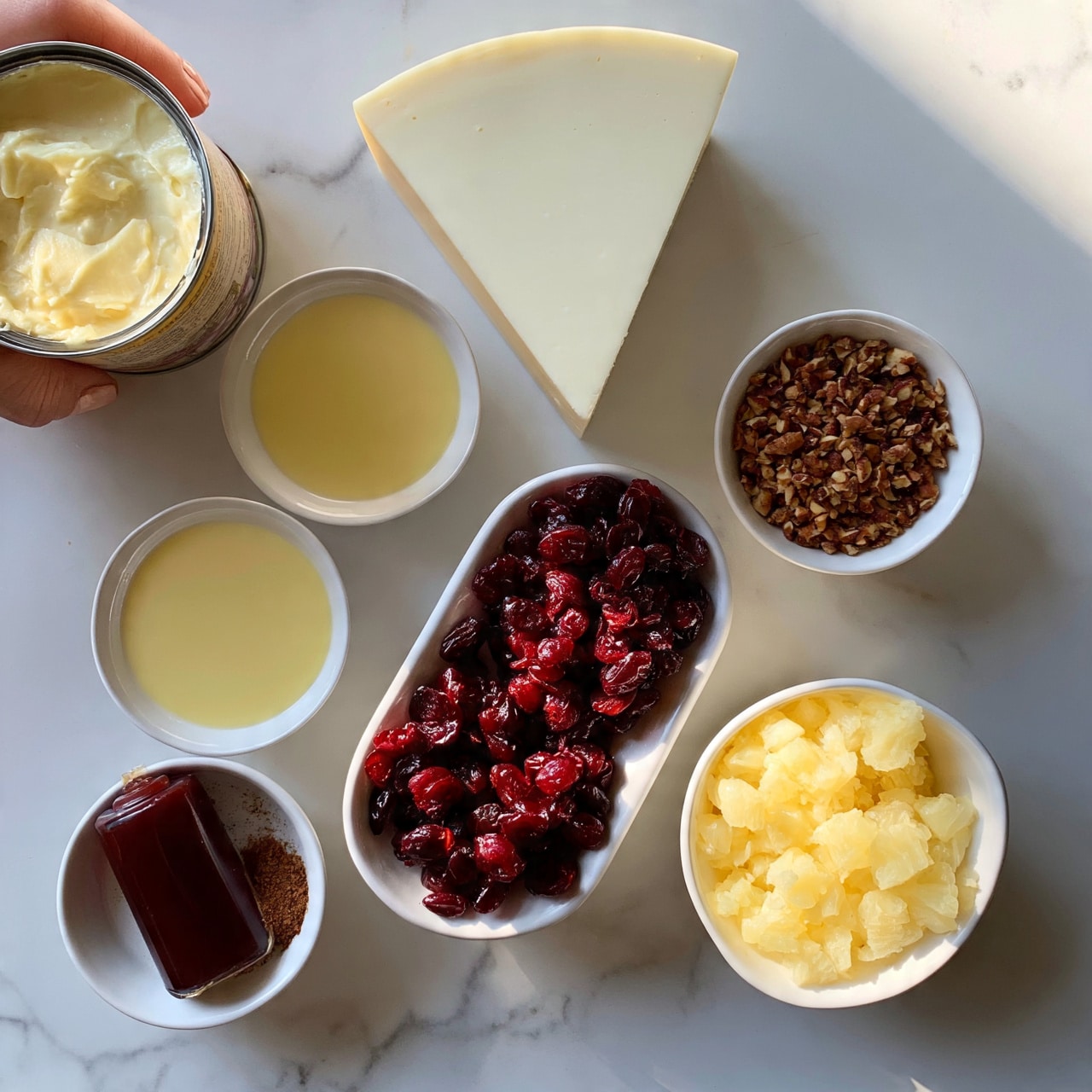 square image The image shows ingredients for a recipe arranged neatly on a white marbled surface. There is one large triangular slice of soft white cheese positioned near the top center. To the left, a woman's hand is holding a can of creamy cheese spread. Below the cheese slice, two small white bowls hold smooth pale yellow liquids. To the right of those, a small white bowl contains finely chopped brown nuts. In the center, a white bowl is filled with deep red dried cherries. Below and slightly right, one white bowl holds light yellow crushed pineapple pieces. To the left, there is a small white bowl with a dark red bottle of flavoring and a small amount of brown spice next to it. The arrangement is clean and organized with soft natural lighting, photo taken with an iphone --ar 4:5 --v 7