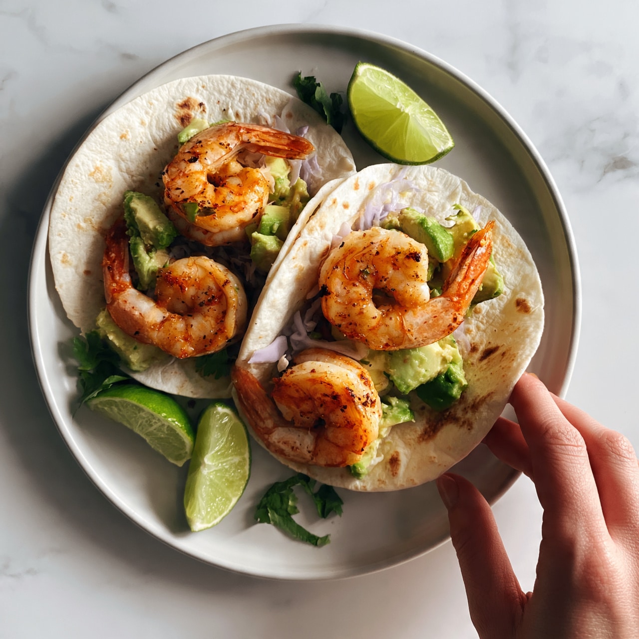 square image The image shows a round white plate holding two soft tacos stacked side by side. Each taco is filled with layers starting with soft, lightly toasted white tortillas as the base. On top of the tortillas is a layer of grilled orange shrimp with slight char marks, followed by chunks of green avocado and small pieces of light purple cabbage for crunch. Bright green lime wedges are placed on the plate, along with some fresh green herbs, adding vibrant color contrast. The plate is set on a white marbled surface, and a woman's hand is reaching toward the tacos. Photo taken with an iphone --ar 4:5 --v 7square imagesquare image