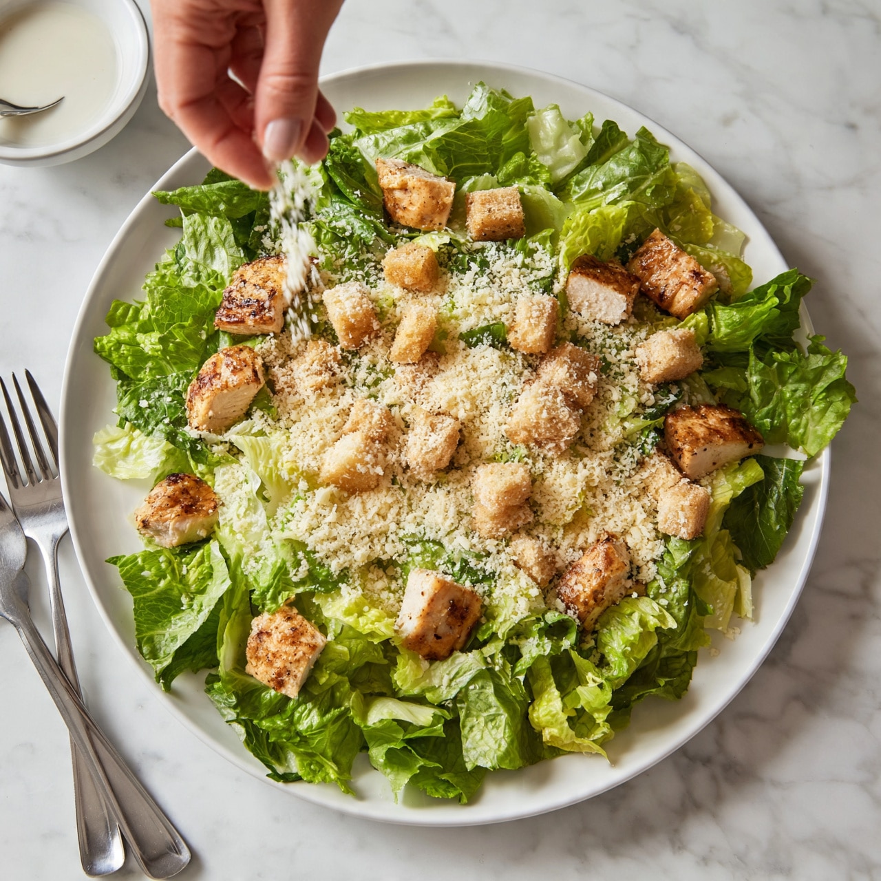 square image A large white plate holds a fresh salad with several layers: the bottom layer is bright green Romaine lettuce leaves, roughly torn; on top are pieces of golden brown grilled chicken scattered evenly; small, crunchy light brown croutons are spread across; a sprinkling of finely grated pale yellow cheese covers the salad; a woman's hand is sprinkling thin white dressing from a small white bowl with a spoon on the side; the plate is placed on a white marbled surface, with two stainless steel forks positioned nearby. photo taken with an iphone --ar 4:5 --v 7
