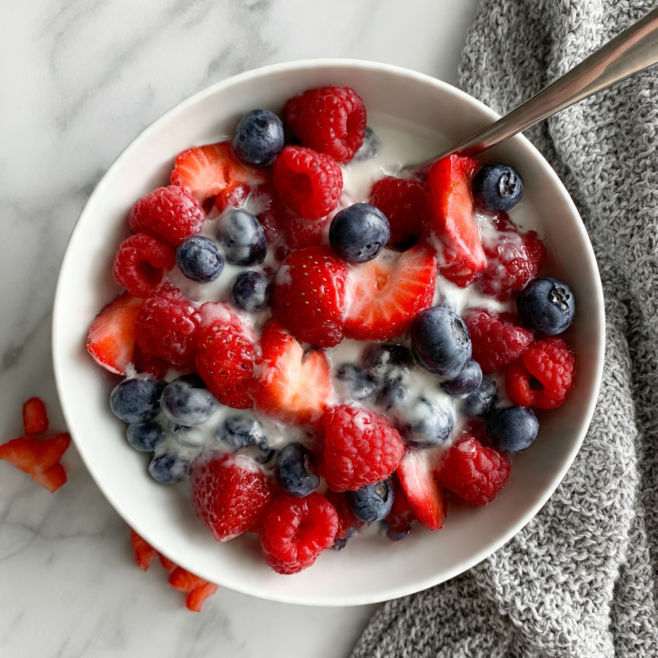 square image A white bowl filled with a colorful mix of fresh berries, including sliced strawberries, whole blueberries, and raspberries. The berries are topped with a light layer of white cream or yogurt, giving a soft contrast to the bright reds and deep blues. The bowl is placed on a white marbled surface, next to a gray knitted cloth and some scattered strawberry pieces. A metallic spoon rests inside the bowl, ready for serving. photo taken with an iphone --ar 4:5 --v 7