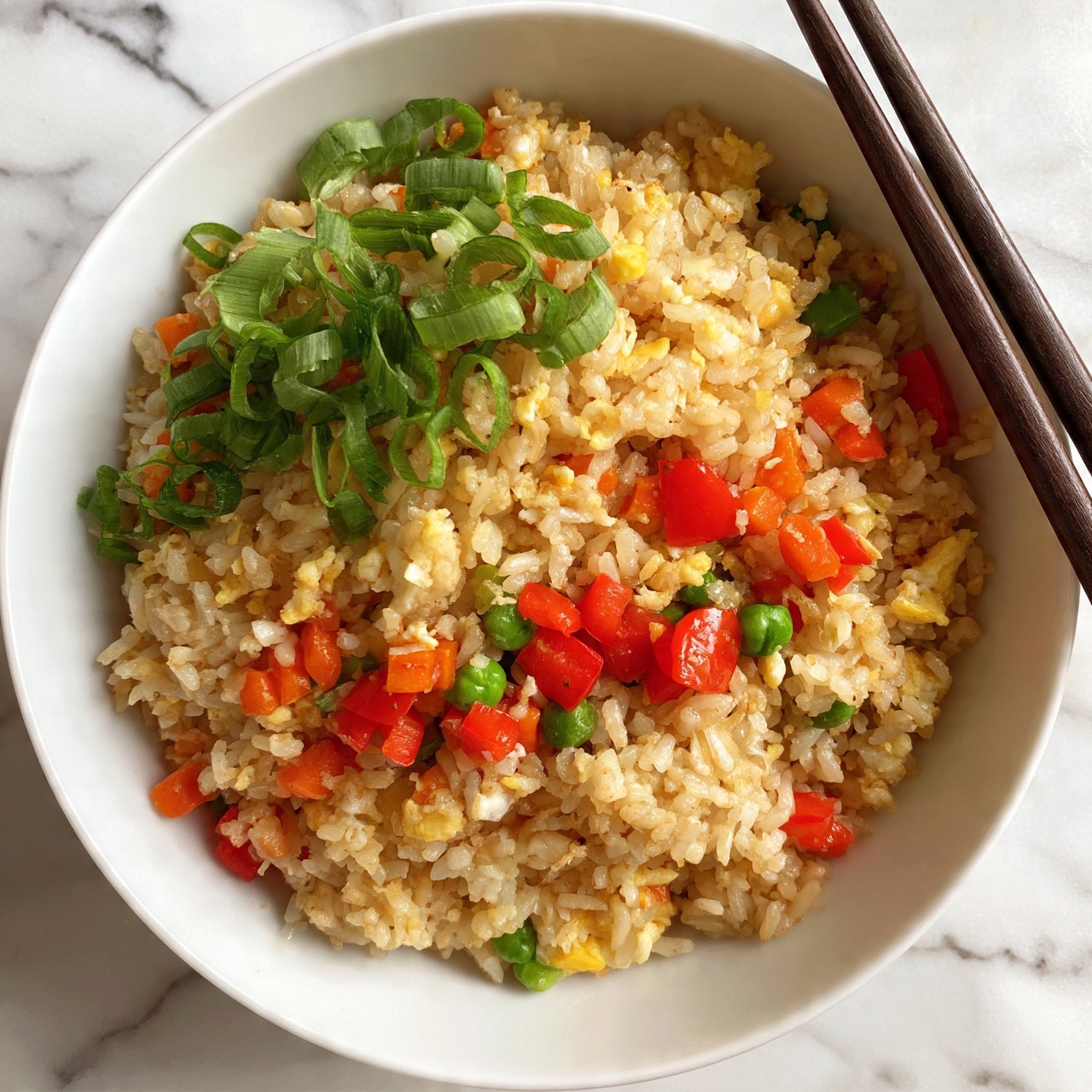 square image A white bowl filled with fried rice showing three main layers: the base layer is light brown rice mixed evenly with small pieces of scrambled eggs and orange carrot bits; the middle layer has bright red diced bell peppers and green peas scattered throughout; the top layer is garnished with fresh, curled green onion slices that add a pop of green color. The bowl sits on a white marbled surface, and a pair of dark wooden chopsticks rests on the right side of the bowl. Photo taken with an iphone --ar 4:5 --v 7