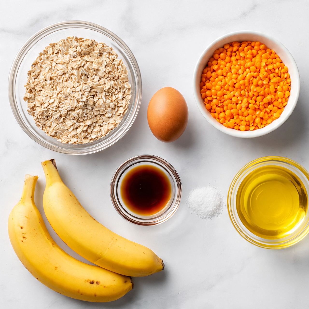 square image The image shows ingredients for a recipe arranged on a white marbled surface. At the bottom left, there are two ripe yellow bananas with small brown spots. Above them to the right, there is a clear glass bowl filled with light brown rolled oats. Next to the oats bowl on the right, an egg is placed directly on the surface. To the right of the egg, a white bowl holds bright orange lentils. Below the oats bowl, a small clear bowl contains a dark amber liquid, likely vanilla extract. Next to it on the right is a tiny pile of white salt on the surface. To the right of that is a clear glass bowl filled with yellow cooking oil. The lighting is bright and natural. photo taken with an iphone --ar 4:5 --v 7