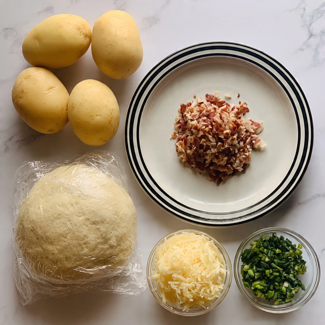 square image The image shows four whole yellow potatoes placed on a white marbled surface in the top left corner. Below the potatoes is a wrapped round dough ball sitting on clear plastic wrap. To the right, a white plate with a thick black rim holds a pile of finely chopped cooked bacon in the center. Below the plate, there are two small clear glass bowls side by side, one filled with shredded pale yellow cheese and the other with chopped green onions. The lighting is soft, giving the items a fresh and natural look. Photo taken with an iphone --ar 4:5 --v 7