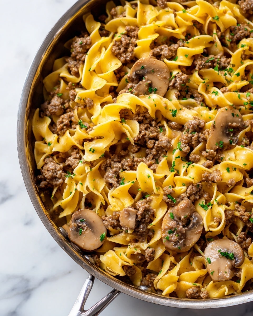 The image shows a close-up of a skillet filled with cooked curly egg noodles mixed with ground meat and sliced mushrooms. The noodles are yellowish with a soft texture, twisting and folding among small pieces of browned meat and light brown mushroom slices. The mixture is coated in a rich brown sauce, which adds a glossy look, and it is sprinkled with small green bits of parsley evenly spread on top, adding color contrast. The skillet itself has a metallic rim, and the background is a white marbled surface. photo taken with an iphone --ar 4:5 --v 7