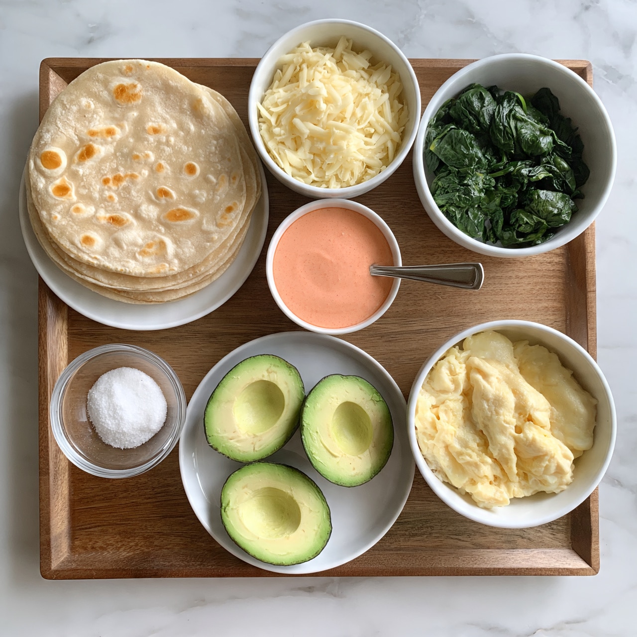 square image The image shows a wooden tray holding seven items arranged neatly for a meal. On the left, a stack of light brown tortillas with golden spots rests on a white plate. Below that, a small clear glass bowl contains white salt. In the middle bottom, a white plate holds three halved avocados with bright green skin and pale green flesh visible. Above the avocados is a small white bowl filled with shredded pale yellow cheese. To the right of the cheese, a white bowl contains cooked dark green spinach. Below the spinach, another white bowl holds light yellow scrambled eggs with a soft texture. In the center, between the avocado and spinach, a small white bowl contains a smooth pink-orange sauce with a spoon in it. All items sit on a white marbled surface. photo taken with an iphone --ar 4:5 --v 7