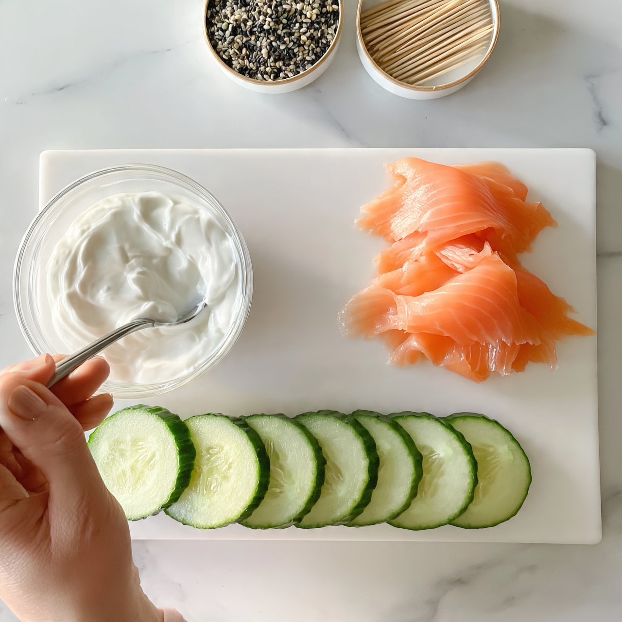square image The image shows a white cutting board on a white marbled surface. On the board, there are three parts: two small piles of thin, smooth salmon slices with a pink-orange color on the right side, a row of several bright green cucumber slices with a crisp texture and visible seeds at the bottom, and a woman's hand holding a spoon stirring a small clear glass bowl of thick white cream on the left. Above the board are two small round containers: one filled with dark and light mixed seeds, the other holding wooden toothpicks. The scene is bright and clean. photo taken with an iphone --ar 4:5 --v 7