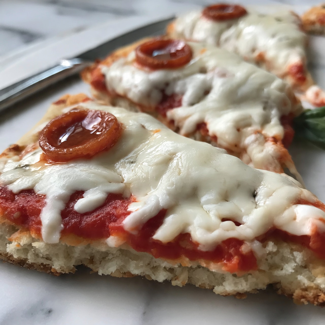 square image The image shows a close-up of a pizza with three visible slices on a white marbled surface. The pizza has three layers: a pale, slightly crispy crust at the bottom with a rough texture, a layer of bright red tomato sauce spread unevenly in the middle, and a thick layer of melted white mozzarella cheese on top that covers most of the pizza, stretching and bubbling in places. Each slice is topped with a single round pepperoni slice, which is glossy and reddish-brown. There is also a small green basil leaf visible on one side of the pizza. A silver knife lies next to the pizza on the white marbled surface. Photo taken with an iphone --ar 4:5 --v 7