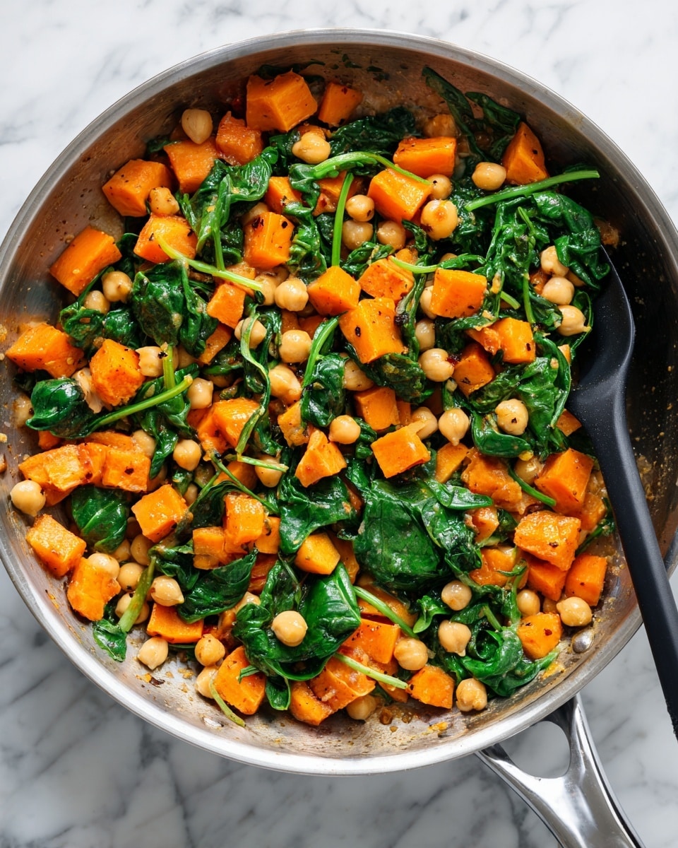 The image shows a close-up of a cooking pan filled with a colorful mix of diced orange sweet potatoes, light beige chickpeas, and fresh dark green spinach leaves. The sweet potatoes and chickpeas are small cubes and round shapes scattered evenly across the pan’s surface, while the spinach leaves are wilted and spread through the mix. The pan sits on a white marbled surface, and a black kitchen spatula is resting inside the pan to the right side. The whole dish looks warm and lightly cooked, with a vibrant and fresh contrast between the bright orange, pale yellow, and deep green colors. Photo taken with an iphone --ar 4:5 --v 7